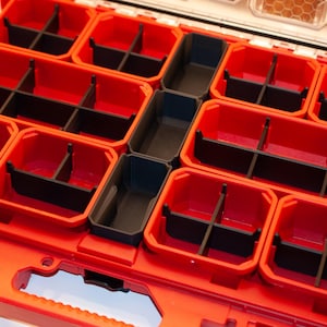 May include: A red and black organizer with multiple compartments. The organizer has a handle and a latch. The compartments are divided by black dividers. The organizer is designed for storing small items.