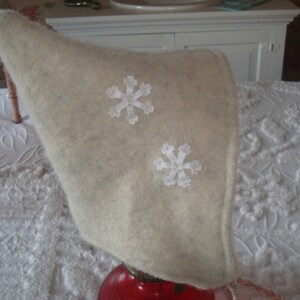 May include: A white felt elf hat with white snowflake embroidery. The hat is on a white bedspread.