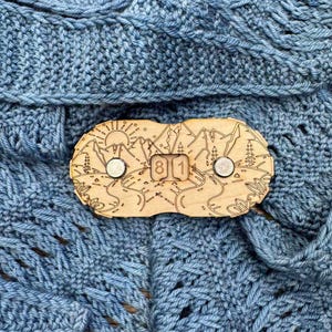 Puede incluir: Un broche de chal de madera con un diseño de montaña y sol cortado con láser, con los números "81". El broche tiene dos cierres magnéticos de color plateado. El broche se muestra sobre una tela de punto azul.