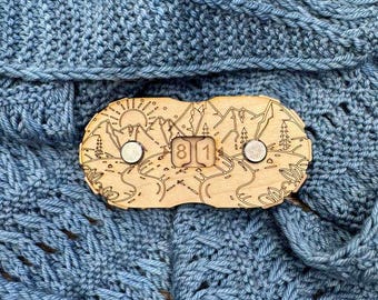 May include: A wooden shawl pin with a laser-cut mountain and sun design, featuring the numbers "81". The pin has two silver-colored magnetic closures. The pin is displayed on a blue knitted fabric.