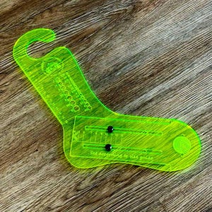 May include: A bright green plastic sock gauge with a circular scale and a sliding gauge. The gauge is labelled "Town Yarn" and has the text "Set according to size guide".
