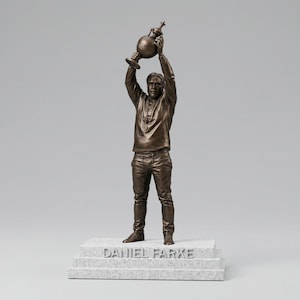 Puede incluir: Estatua de bronce de un hombre sosteniendo un trofeo por encima de su cabeza. La estatua está sobre una base de piedra con la inscripción "DANIEL FARKE". El hombre lleva una camisa de manga larga, pantalones y una medalla.