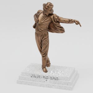 Könnte beinhalten: Bronzefarbene Statue einer tanzenden Figur mit Hut und Jacke, ein Arm ausgestreckt. Die Statue steht auf einem weißen, mehrschichtigen Sockel mit der Aufschrift "KING OF POP". Die Figur befindet sich in einer dynamischen Pose.