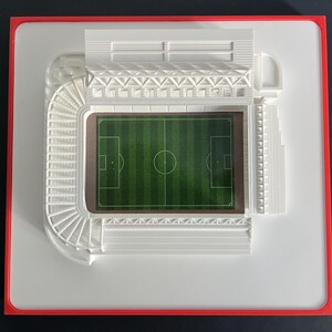 May include: A miniature stadium model with a green field and white seating. The model is set on a white base with a red border. The stadium features detailed architectural elements.