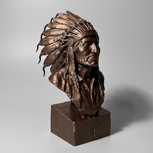 May include: A bronze-coloured bust sculpture depicting a person wearing a traditional feathered headdress. The figure has braided hair, a collared shirt, and a stoic expression. The sculpture is mounted on a solid, rectangular bronze-coloured base.