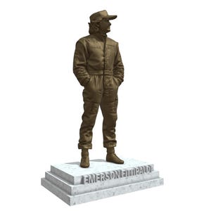 May include: A bronze-colored statue of a person in a racing suit and cap, standing on a marble base. The base is inscribed with the name "EMERSON FITTIPALDI". The statue has its hands in its pockets.