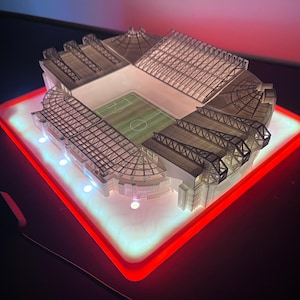 May include: A miniature stadium model with a green field and detailed architectural features. The model sits on a glowing red and white illuminated base, creating a striking visual effect. The stadium's roof and seating areas are intricately designed.