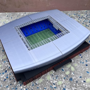 May include: A miniature stadium model with a silver roof, blue seating, and a green field. The model has a rectangular shape with a black base and red trim. The field has white markings.