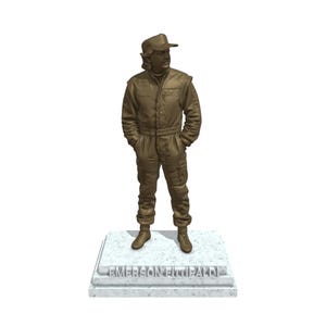 May include: A bronze-colored statue of a person in a racing suit and cap, standing with hands in pockets. The statue is mounted on a rectangular base with the name "EMERSON FITTIPALDI" engraved on it.