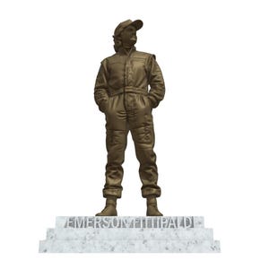 May include: A bronze statue of a person in a racing suit and cap, standing on a marble base. The statue has its hands in its pockets and is facing forward. The name "EMERSON FITTIPALDI" is inscribed on the base.