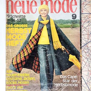 May include: Vintage fashion magazine cover featuring a woman in a yellow hat, scarf, and top, with a dark cape lined with plaid. The magazine title is "Neue Mode" with text in German. The cover also includes the text "Das Cape: Star der Herbstmode."