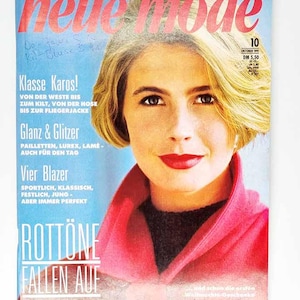 Puede incluir: Una portada de revista con una mujer de pelo rubio y labios rojos. El título de la revista "neue mode" está en rojo, con texto en alemán. La portada presenta un abrigo rosa y un fondo azul, con texto promocional.