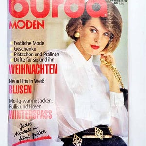 May include: A vintage magazine cover with the title "Burda Moden" in red. The cover features a woman with short hair and red lipstick. Text includes "WEIHNACHTEN" and "BLUSEN". The woman is wearing a white blouse and a black belt with gold detailing.