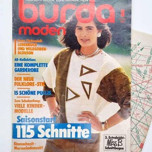 May include: A vintage Burda Moden magazine from August 1985, issue M 2017 E, featuring a cover model wearing a white and brown top with triangular cutouts. The cover includes text such as 115 Schnitte and Miss B Schnittbogen, alongside a sewing pattern sheet.