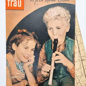 May include: Vintage magazine cover featuring two children. A boy plays a recorder, while a girl smiles. The cover has text in German, including "Stimme der Frau" and "Mode Heim Leben." The magazine is aged with a worn appearance.