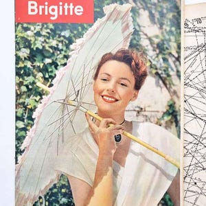 以下が含まれることがあります： 赤いボックスに「Brigitte」という文字が入ったヴィンテージ雑誌の表紙。表紙には、白いとピンクの日傘を持った笑顔の女性が写っています。背景には緑と地図が含まれています。