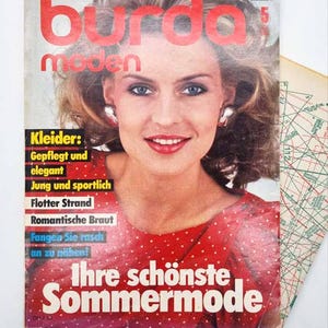 May include: A vintage magazine cover featuring a woman with blonde hair and red lipstick. The magazine title "Burda Moden" is in large red letters. Text on the cover includes "Ihre schönste Sommermode" and other German phrases.