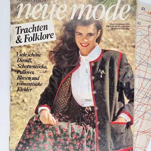 May include: A vintage magazine cover featuring a woman in traditional German attire. The cover text reads "neue mode" and "Trachten & Folklore." The woman wears a dark jacket with red trim, a white blouse, and a floral skirt. The magazine is printed in Germany.
