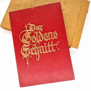 Pode incluir: Um livro vermelho com letras douradas e uma capa bege. As letras douradas dizem "Der Goldene Schnitt". O livro está aberto, revelando as páginas internas. A capa está ligeiramente aberta.