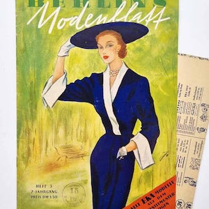 May include: Vintage magazine cover titled "Berlins Modenblatt" featuring a woman in a blue dress and hat. The cover has green and yellow tones with text in German. Additional paper with text is visible.