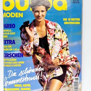 Puede incluir: Una portada de revista con una mujer que lleva una chaqueta de estampado floral y pantalones cortos negros. La revista se titula "Burda Moden" y el texto de la portada dice "Weltweit in Mode" y "Die 10 besten Obstkuchen".