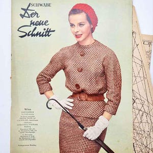 May include: Vintage fashion magazine cover featuring a woman in a brown tweed suit, red hat, and white gloves. The magazine cover has the text "Der neue Schnitt" and the date "9/1958". The woman is holding a black umbrella.