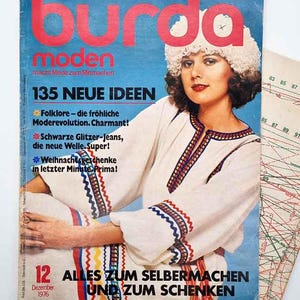 May include: Vintage Burda Moden magazine cover featuring a woman in a white embroidered outfit and a fur hat. The cover has text in German, including "135 NEUE IDEEN" and "Dezember 1976". A pattern sheet is visible.