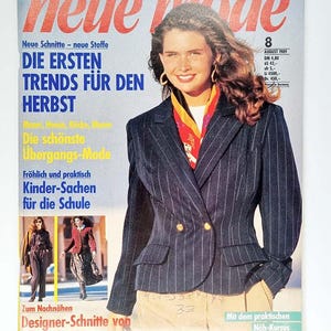 Puede incluir: Una portada de revista de moda con una mujer con una blazer de raya diplomática azul marino con botones dorados, una bufanda colorida y pantalones beige. El título de la revista es "neue mode" con texto en alemán, incluyendo "Die ersten Trends für den Herbst".