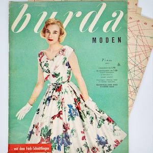 Può includere: Una copertina di rivista vintage con una donna in un abito bianco con stampa floreale. La rivista è intitolata "burda MODEN" e la data è "7/1956 JULI". Il testo "... mit dem Farb-Schnittbogen" si trova nella parte inferiore della copertina.