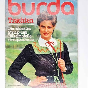 Könnte beinhalten: Ein Magazin-Cover mit dem Wort "burda" in Rot. Das Model trägt eine schwarze Strickjacke mit grünen und orangenen Details, ein weißes Hemd mit Kragen und eine rote Schleife. Der Magazintitel ist "Trachten" mit deutschem Text darunter.