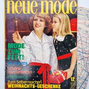 Pode incluir: Uma capa de revista vintage com o título "neue mode" em amarelo. Mostra duas mulheres, uma com uma faísca e a outra segurando um presente. O texto inclui "MODE ZUM FEST!" e "WEIHNACHTS-GESCHENKE".