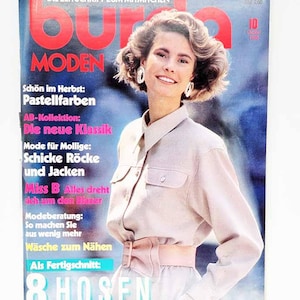 Puede incluir: Una portada de revista con una mujer vestida con un atuendo rosa claro. La portada presenta el texto "BURDA MODEN" en letras rojas grandes. Otros textos incluyen "DIE ZEITSCHRIFT ZUM MITMACHEN" y "8 HOSEN". El fondo es una mezcla de azul y blanco.