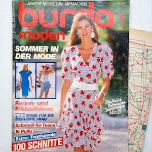 Può includere: Una copertina di rivista di moda vintage con una donna in un abito bianco con motivi floreali rossi. La rivista è intitolata "Burda Moden" in grandi lettere rosse e include testo in tedesco. Sono visibili anche fogli di modelli.