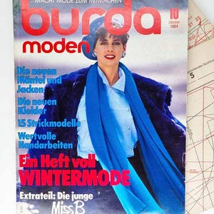 May include: A vintage Burda Moden magazine cover from October 1984, featuring a woman in a blue outfit and a blue scarf. The cover has red text and German text, with a pattern sheet visible in the background. The magazine is focused on winter fashion.