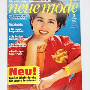 May include: A magazine cover featuring a woman wearing a red jacket and a yellow watch. The magazine is titled "neue mode" and has the text "Mit dem großen Handarbeitsteil" and "Hier sind sie:" on the cover. The magazine cover also has the text "Neu! Großer Schnitt-Service für unsere Leserinnen".