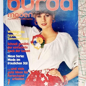 May include: A vintage Burda Moden magazine cover featuring a woman in a white blouse and a red floral skirt. The magazine title is in large red letters. Text on the cover includes phrases in German about summer blouses and sewing.