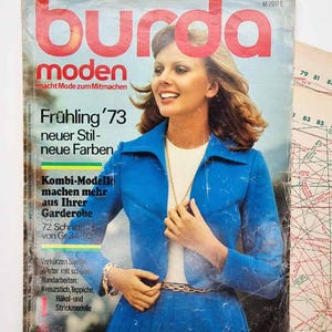 以下が含まれることがあります： 1973年のBurda Moden誌の表紙。青いスーツを着た女性が特徴です。雑誌のタイトルは赤い大きな文字で、ドイツ語のテキストが添えられています。表紙は少し古びています。