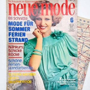 Pode incluir: Uma edição vintage da revista Neue Mode de junho de 1975 com uma capa que mostra uma pessoa vestindo uma blusa turquesa brilhante com um detalhe floral no ombro. A capa inclui texto em alemão e é exibida ao lado de um molde de costura.