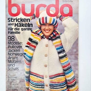 May include: A Burda magazine cover titled Stricken und Häkeln für die ganze Familie, featuring a person wearing a cream-coloured knit cardigan with wooden buttons and a matching hat and scarf with horizontal stripes in red, blue, yellow, and black.