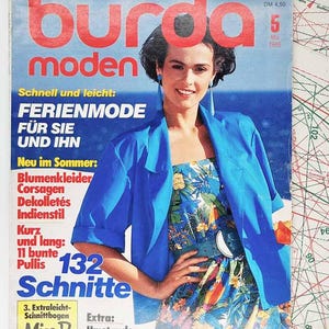 May include: A vintage Burda Moden magazine cover featuring a woman in a blue jacket and floral dress. The magazine cover has text in German, including "Ferienmode" and "132 Schnitte". The background includes sewing patterns.