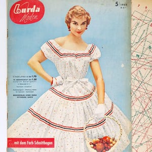 Könnte beinhalten: Ein Vintage-Magazincover mit einer Frau in einem weißen Kleid mit roten und blauen Verzierungen, die einen Korb mit Obst hält. Das Magazin heißt "Burda Moden" und das Datum ist Mai 1955.
