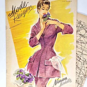 Op de afbeelding: Vintage mode tijdschriftcover met een vrouw in een paars pak die bloemen vasthoudt. De cover heeft de tekst "Mode-Reigen" en "Nimon Modelle" in sierlijke letters. Een patroonvel is zichtbaar op de achtergrond.