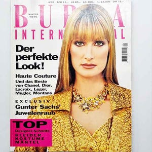 Może przedstawiać: Okładka magazynu z blondynką w złotej kurtce i naszyjniku. Magazyn to "BURDA INTERNATIONAL" z tekstem "Der perfekte Look!" i "Haute Couture". Różowy prostokąt na dole zawiera napis "TOP Designer-Schnitte".