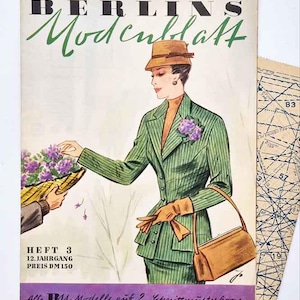 May include: A vintage magazine cover with the title "BERLINS Modenblatt" in German, featuring a woman in a green pinstripe suit, hat, and gloves. She holds a brown handbag. A basket of purple flowers is offered. The magazine also has German text.