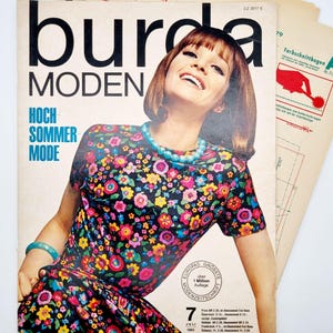 May include: A vintage Burda Moden magazine cover featuring a woman wearing a colorful floral dress with a turquoise necklace. The cover text reads "Burda Moden" and "Hoch Sommer Mode".
