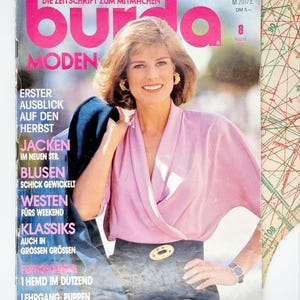 Puede incluir: Una portada de revista vintage con una mujer con una blusa rosa y una falda oscura. El título de la revista "Burda Moden" está en rosa. La portada incluye texto en alemán y un patrón es visible en el fondo.