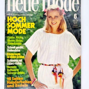 Könnte beinhalten: Ein Magazincover mit einer Frau, die ein weißes Oberteil mit einem bunten Gürtel und einer Blumenkrone trägt. Das Magazin heißt "neue mode" und der Titeltext lautet "Hoch Sommer Mode".