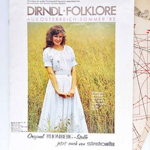 Puede incluir: Una portada de revista vintage con una mujer con un vestido floral blanco y azul, de pie en un campo. La portada dice "DIRNDL FOLKLORE AUS ÖSTERREICH SOMMER '85" y "Original RHOMBERG-Stoffe".