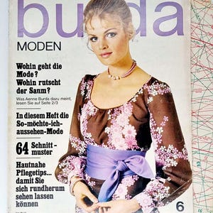 Puede incluir: Una portada de revista con una mujer que lleva un vestido floral marrón y rosa con una faja morada. La revista se titula "buda MODEN" y el texto "64 Schnitt-muster" es visible. La portada de la revista es de junio de 1970.