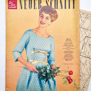 May include: Vintage magazine cover featuring a woman in a blue patterned dress holding red roses. The magazine title reads "NEUER SCHNITT" with additional text and a pattern sheet visible. The cover has a retro aesthetic.
